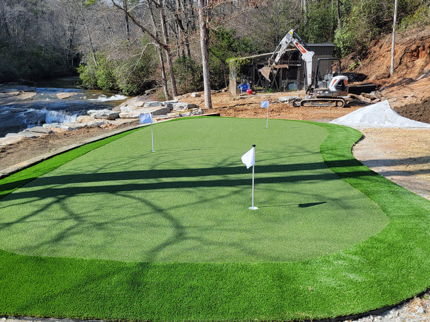 Synthetic turf putting green by Synthetic Turf Concepts, elegantly set beside a beautiful body of water and rocky landscape, merging sport with nature.